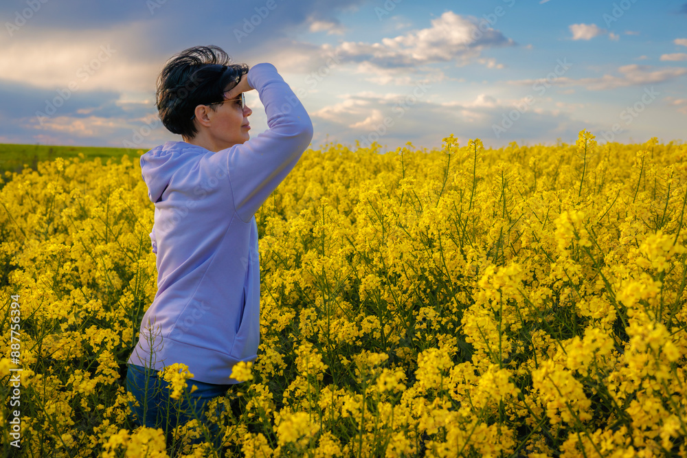 A woman standing in a rapeseed field and looking into the distance with her hand on her head like a visor. A woman standing in a rapeseed field and looking into the distance with her hand on her head like a visor.