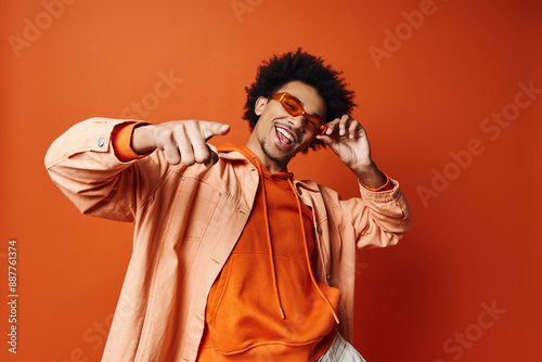 Bild auf Leinwand A stylish young African American man with curly hair wearing an orange shirt and sunglasses, posing energetically against an orange background