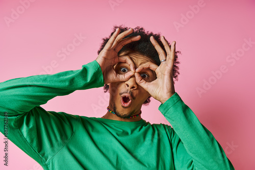 Fotografie A young African American man in casual wear on a pink background, making a playful and expressive face using his hands