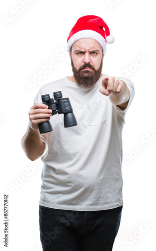 Young caucasian hipster man wearing christmas hat looking though binoculars over isolated background pointing with finger to the camera and to you, hand sign, positive and confident gesture