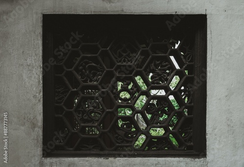 A black hexagonal window with intricate carving, set in a white wall, offers a glimpse of green foliage outside.