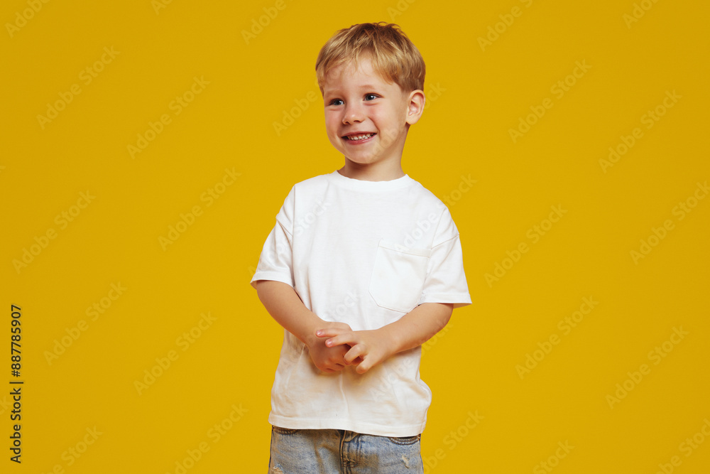 Adorable little blonde cute kid boy wearing white t-shirt, smiling broadly while looking away. Innocent child standing isolated against yellow background. Adorable little blonde cute kid boy wearing white t-shirt, smiling broadly while looking away. Innocent child standing isolated against yellow background.