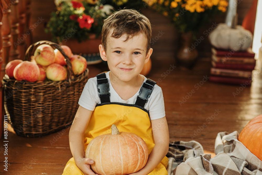 Fototapeta premium Portrait of a Caucasian boy in a yellow jumpsuit sitting on the porch of his house decorated with pumpkins in the autumn season.