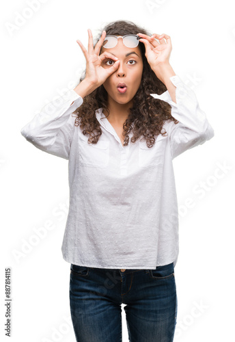 Beautiful young hispanic woman doing ok gesture shocked with surprised face, eye looking through fingers. Unbelieving expression.