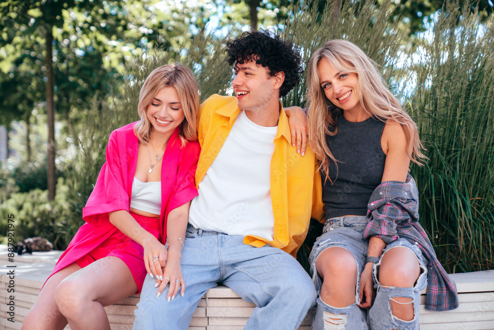 Fototapeta premium Group of young three stylish friends posing in the street. Fashion man and two cute female dressed in casual summer clothes. Smiling models having fun. Cheerful women and guy outdoors