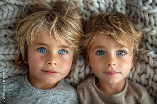 Two young children with striking blue eyes lie on a knitted blanket, capturing a moment of innocence and closeness. Focus on faces, highlighting their expressions. © Dacha AI