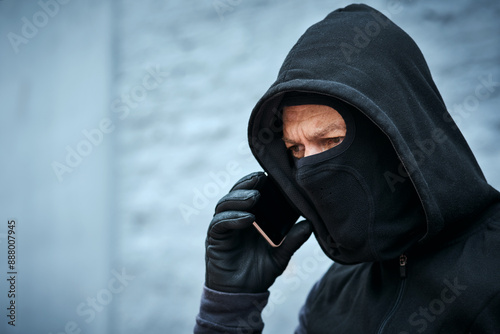Mask, thief or man with phone call for talking, provide instructions and demand ransom for complete mission. Outdoor, male criminal and mobile with face cover for identity protection, hide and fraud