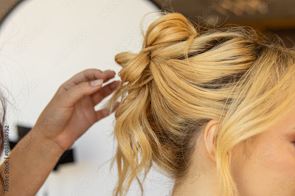 Fototapeta premium Professional stylist styles the hair of blonde woman. Styling hairstyles for wedding or event. High bun of curly hair