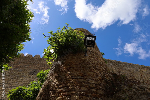 A stone fortress with a lantern is covered with greenery that has sprouted in the wall; blue sky; green tree; stone; bottom-up view; historical  photo