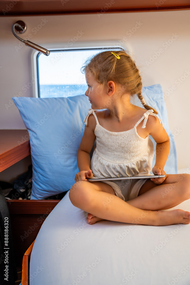 Little girl sits with tablet in the cabin of a yacht and looks out the window during a sea crossing. Yacht life concept Little girl sits with tablet in the cabin of a yacht and looks out the window during a sea crossing. Yacht life concept