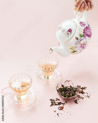 Fancy tea pot pouring rose tea into glass tea cups on pink background 