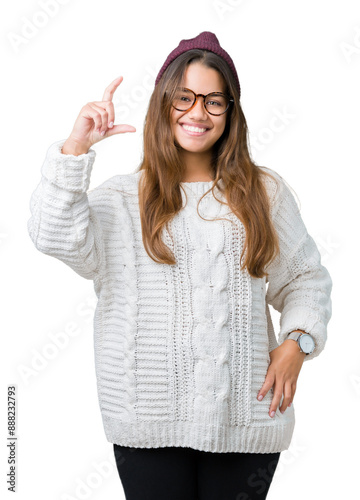 Young beautiful brunette hipster woman wearing glasses and winter hat over isolated background smiling and confident gesturing with hand doing size sign with fingers while looking and the camera