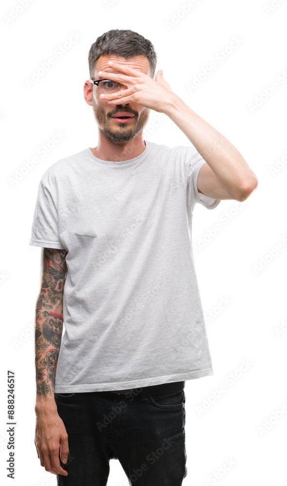 Young tattooed adult man peeking in shock covering face and eyes with hand, looking through fingers with embarrassed expression. Young tattooed adult man peeking in shock covering face and eyes with hand, looking through fingers with embarrassed expression.