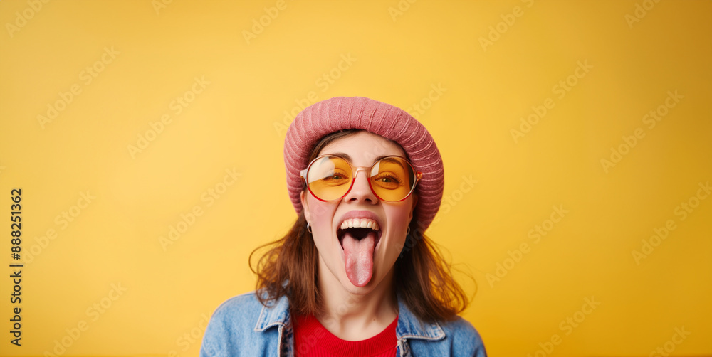A playful woman wearing a beanie and yellow glasses, with her tongue out against a yellow background A playful woman wearing a beanie and yellow glasses, with her tongue out against a yellow background
