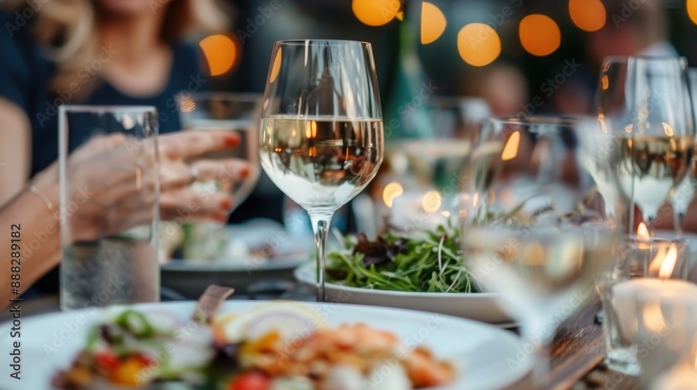 Close-up view of a table set at a social gathering, featuring glasses of wine, fresh salad, and other dishes with several people enjoying the meal in the background. Close-up view of a table set at a social gathering, featuring glasses of wine, fresh salad, and other dishes with several people enjoying the meal in the background.