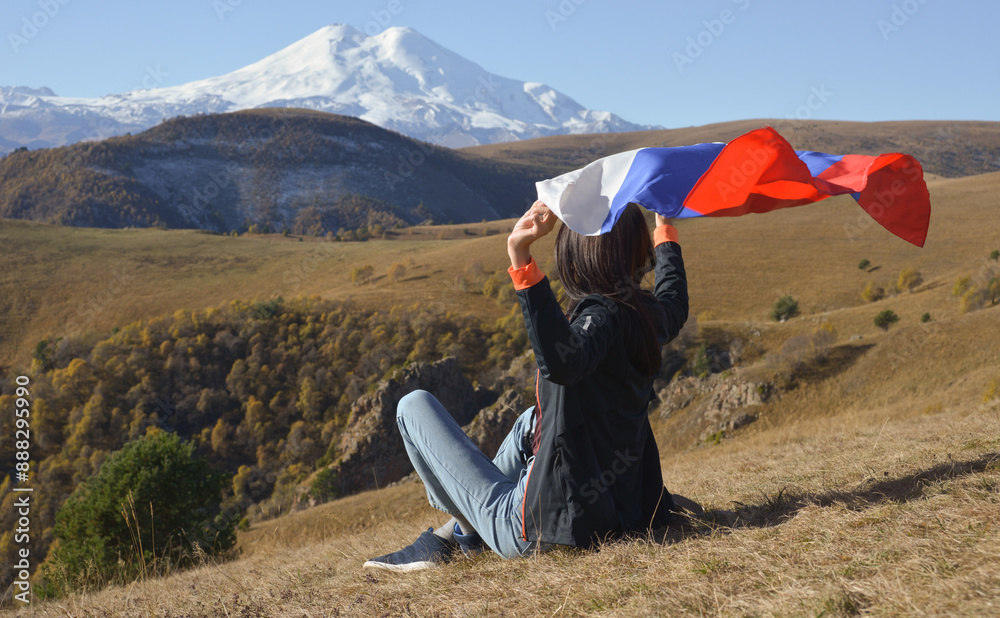 A young brunette woman sits on a stone and looks at the Caucasus Mountains around, a Russian flag flutters in her hands. Tricolor against the backdrop of snow-capped Mount Elbrus