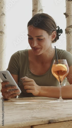 young astonished woman sitting in a cafe using her smartphone to browse the internet, text, chat with friends, or check social media news, while drinking a glass of aperitif and smiling at the camera