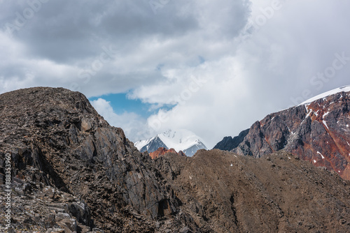 Aerial alpine view to sunlit sheer crags and multicolor mountains against snowy mountain top of dome shape in low clouds. Colorful sharp rocky wall against high snow-white peak far away in cloudy sky. photo