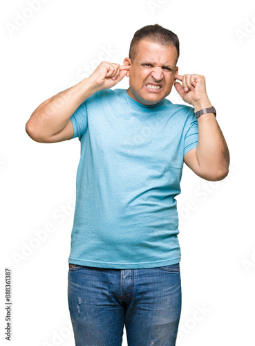 Middle age arab man wearing blue t-shirt over isolated background covering ears with fingers with annoyed expression for the noise of loud music. Deaf concept.