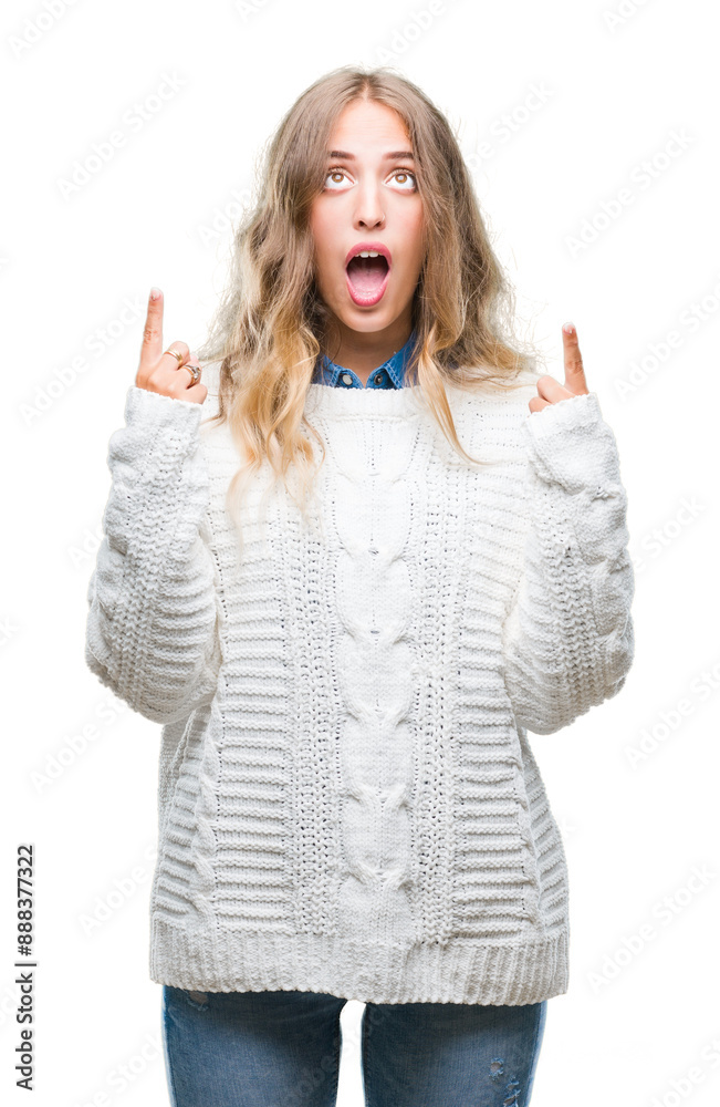 Beautiful young blonde woman wearing winter sweater over isolated background amazed and surprised looking up and pointing with fingers and raised arms.