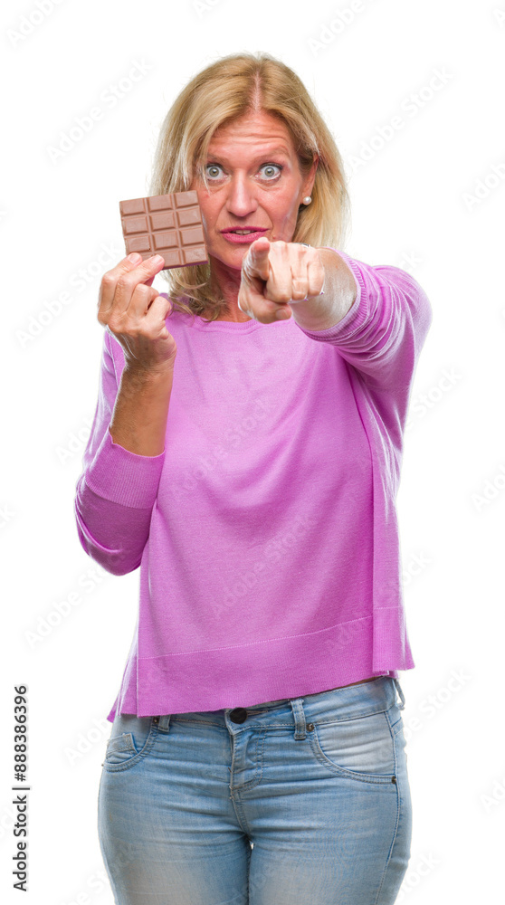 Middle age blonde woman eating chocolate bar over isolated background pointing with finger to the camera and to you, hand sign, positive and confident gesture from the front