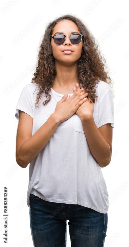 Young hispanic woman wearing sunglasses smiling with hands on chest with closed eyes and grateful gesture on face. Health concept. Young hispanic woman wearing sunglasses smiling with hands on chest with closed eyes and grateful gesture on face. Health concept.