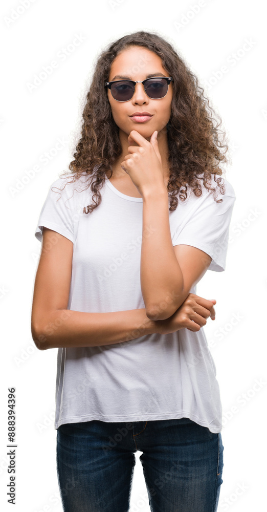 Young hispanic woman wearing sunglasses looking confident at the camera with smile with crossed arms and hand raised on chin. Thinking positive. Young hispanic woman wearing sunglasses looking confident at the camera with smile with crossed arms and hand raised on chin. Thinking positive.