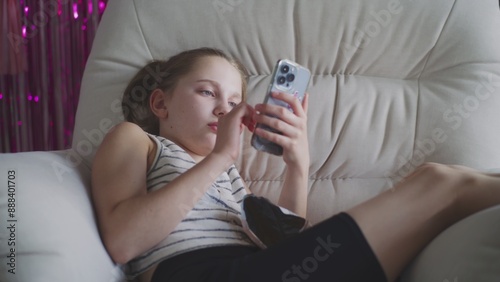 Caucasian teenager uses her mobile phone to surf Internet and chat with friends in social network. Young girl lies on big cozy armchair and rests. Attractive teen spends time in comfort room at home.