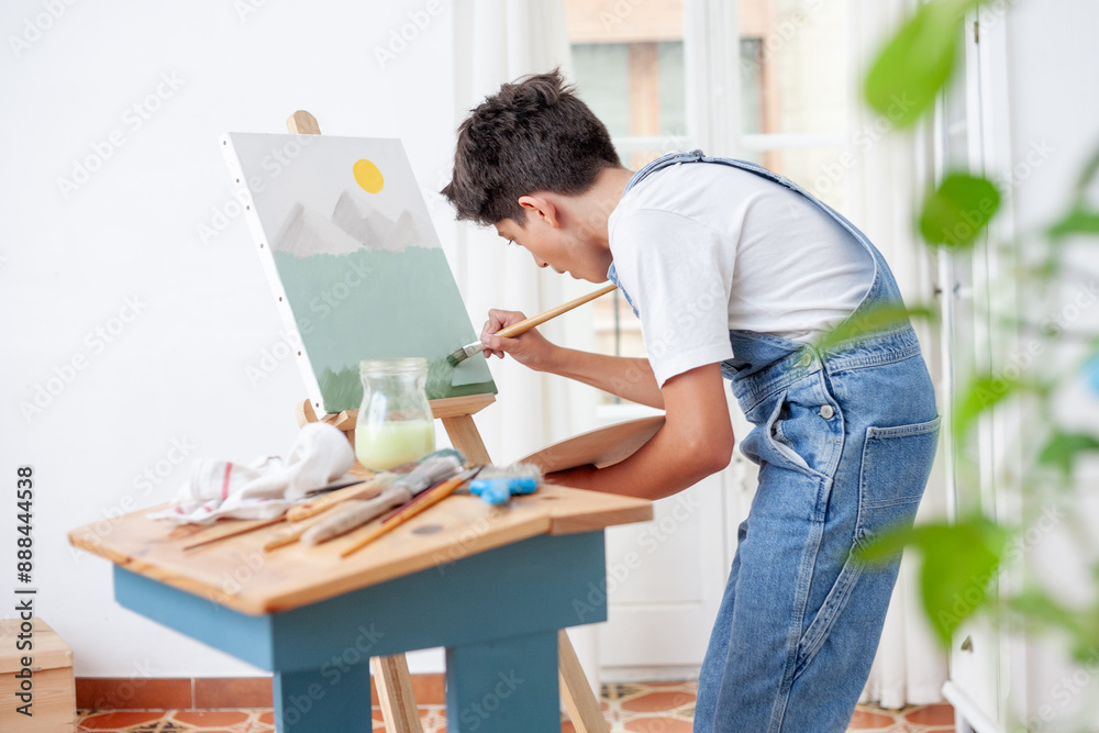 A teenager is painting a landscape with a paintbrush on a canvas on an easel in a living room with plants . A teenager is painting a landscape with a paintbrush on a canvas on an easel in a living room with plants .