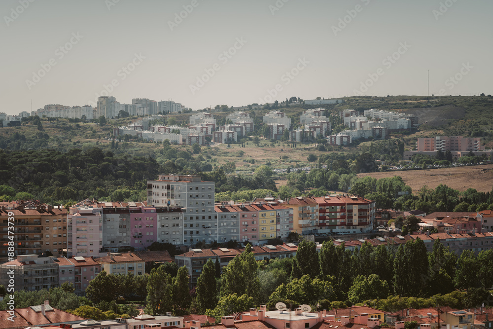 Obraz premium A suburban area near Lisbon featuring colorful residential buildings in the foreground and densely packed apartment blocks on a hillside in the background, surrounded by lush greenery and a clear sky