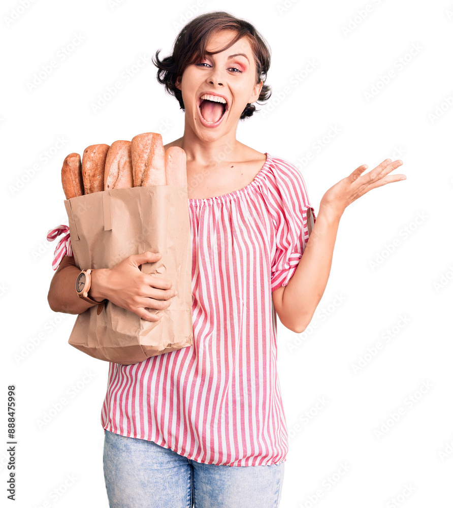 Beautiful young woman with short hair holding paper bag with bread celebrating victory with happy smile and winner expression with raised hands Beautiful young woman with short hair holding paper bag with bread celebrating victory with happy smile and winner expression with raised hands