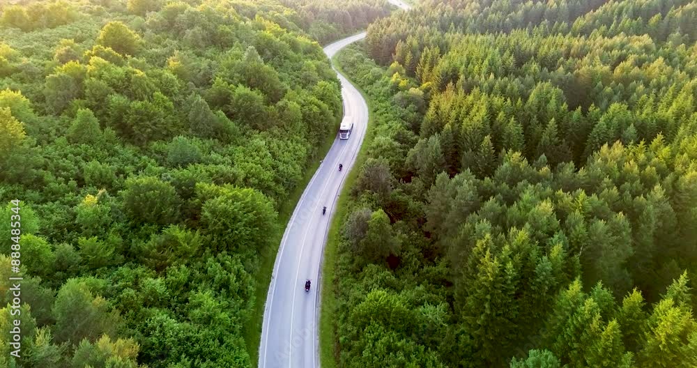 At sunset, three motorcyclists ride along a winding road through a lush, green forest, captured from an aerial view, highlighting their journey under the warm hues of the setting sun