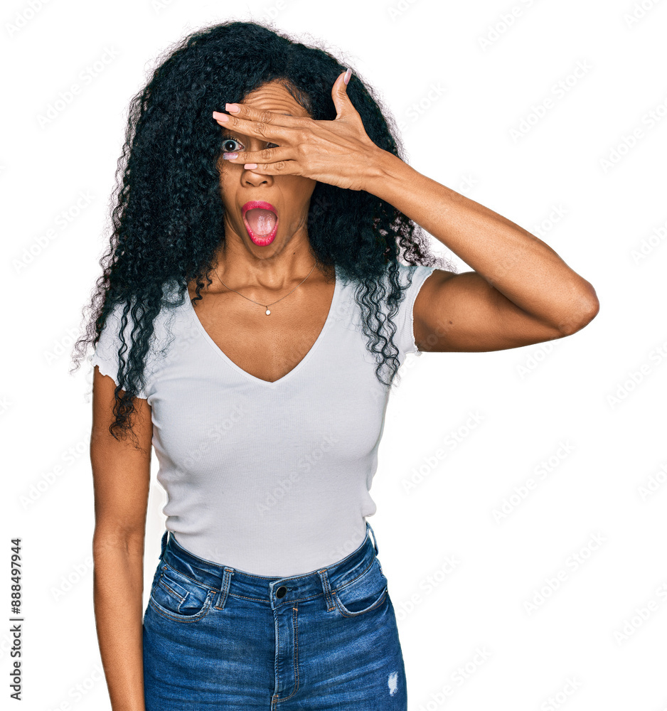 Middle age african american woman wearing casual white t shirt peeking in shock covering face and eyes with hand, looking through fingers with embarrassed expression. Middle age african american woman wearing casual white t shirt peeking in shock covering face and eyes with hand, looking through fingers with embarrassed expression.