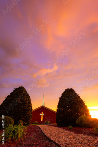 Church with steeple and glowing cross between two large shrubs with pink sky sunset photo