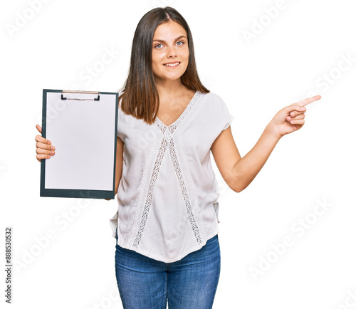 Young caucasian woman holding clipboard with blank space smiling happy pointing with hand and finger to the side