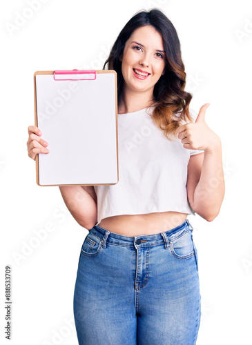 Beautiful young brunette woman holding clipboard with blank space smiling happy and positive, thumb up doing excellent and approval sign