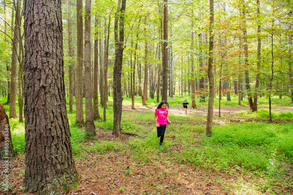 A boy and girl run through a bright green wooded area with streams of morning light in Historic Occoneechee Speedway Trail, Hillsboro, North Carolina A boy and girl run through a bright green wooded area with streams of morning light in Historic Occoneechee Speedway Trail, Hillsboro, North Carolina