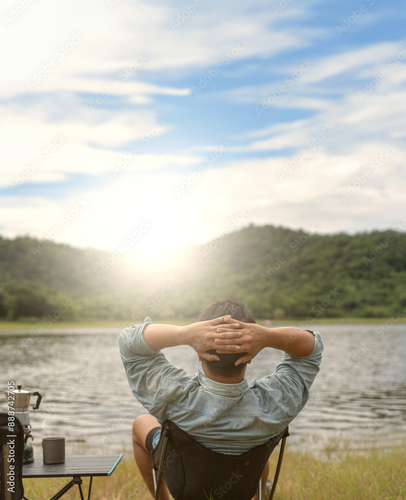 a man sitting on a chair , hands behind their head, relaxing and enjoying the beautiful nature. The background includes a calm lake, lush green hills, and sunny sky with some clouds. The overall mood a man sitting on a chair , hands behind their head, relaxing and enjoying the beautiful nature. The background includes a calm lake, lush green hills, and sunny sky with some clouds. The overall mood