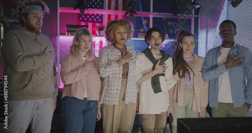 Diverse group of sports fans young people singing anthem watching sports on TV in evening with flags of European Union and United States of America in background