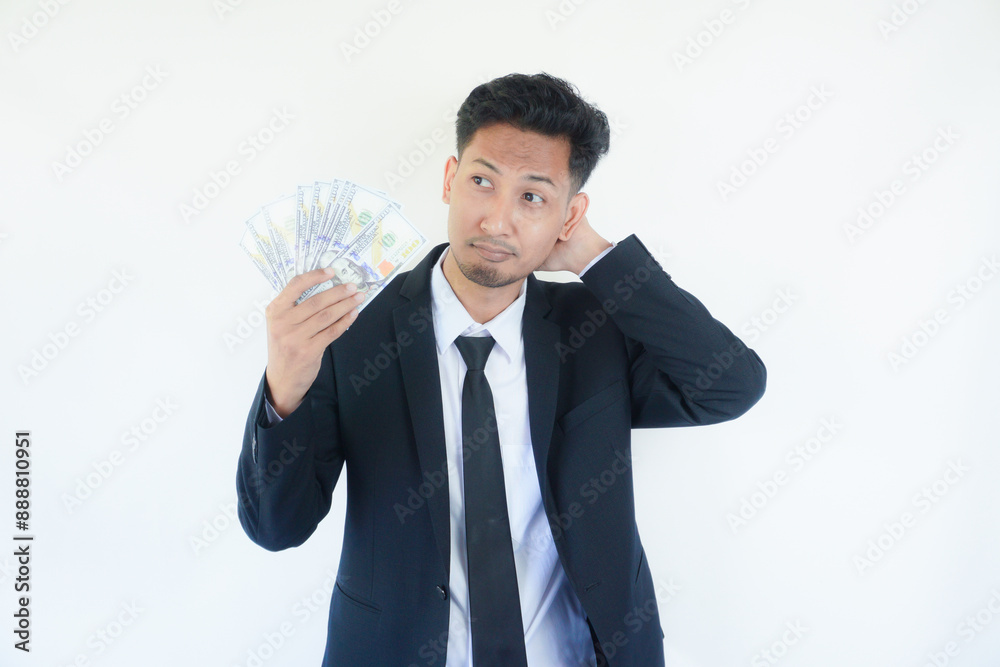 Young Asian man holding dollar bills in studio touching back of head, thinking and making a choice. Young Asian man holding dollar bills in studio touching back of head, thinking and making a choice.
