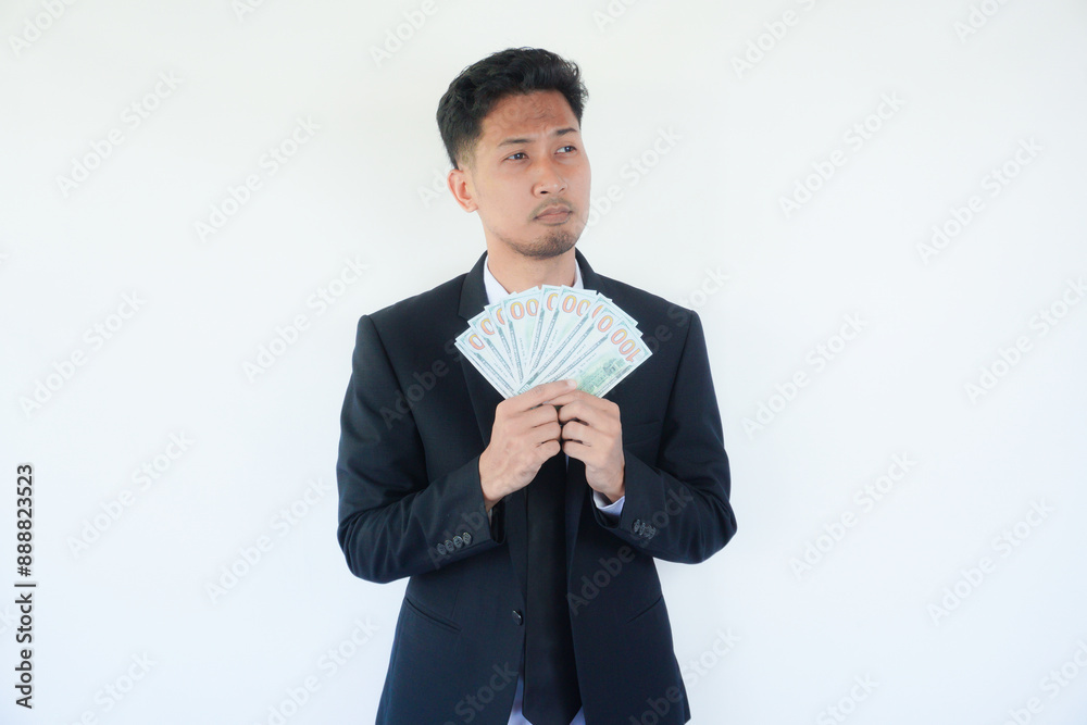 Young Asian man holding dollar bills in studio touching back of head, thinking and making a choice. Young Asian man holding dollar bills in studio touching back of head, thinking and making a choice.