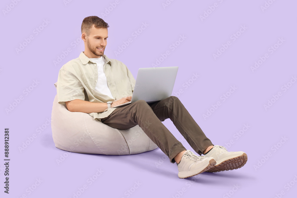 Handsome man using laptop on beanbag against lilac background Handsome man using laptop on beanbag against lilac background