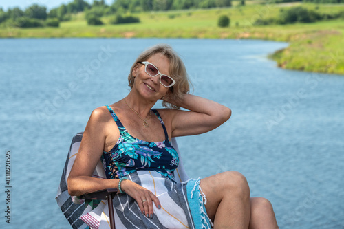 An mature woman in a swimsuit sits in a sun lounger on the river bank.
