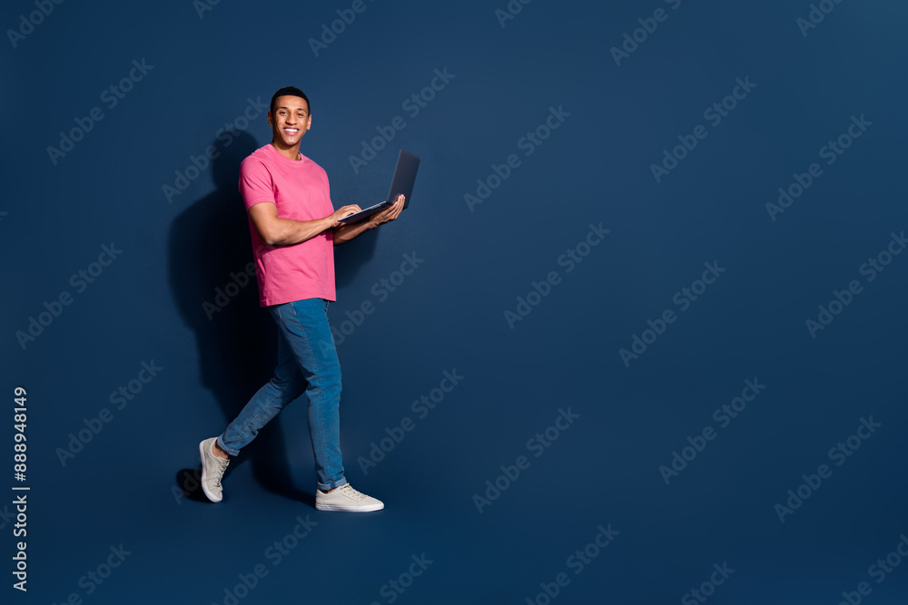Full body length photo of young man in pink t shirt and jeans walking using computer working remote isolated on dark blue color background Full body length photo of young man in pink t shirt and jeans walking using computer working remote isolated on dark blue color background