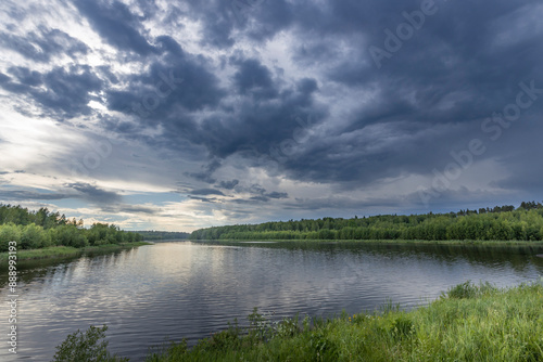 A scenic view of a tranquil river under a dramatic summer sky, with thick, dark clouds gathering overhead and a line of lush green trees along the banks.