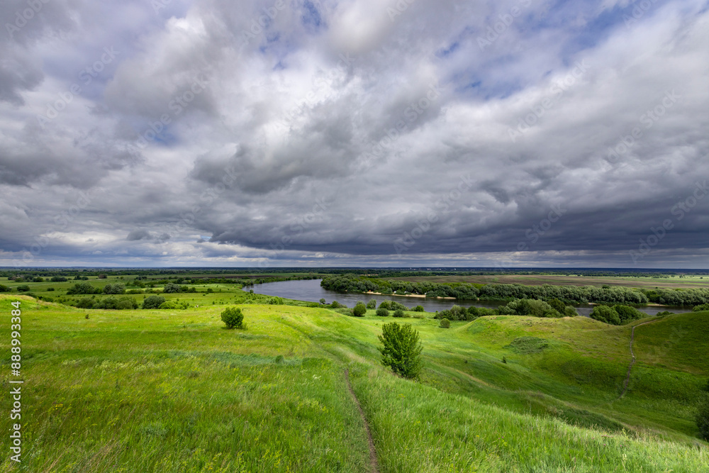 A panoramic view of a winding path leading towards a river amidst rolling green hills, all under a captivating, cloudy sky.