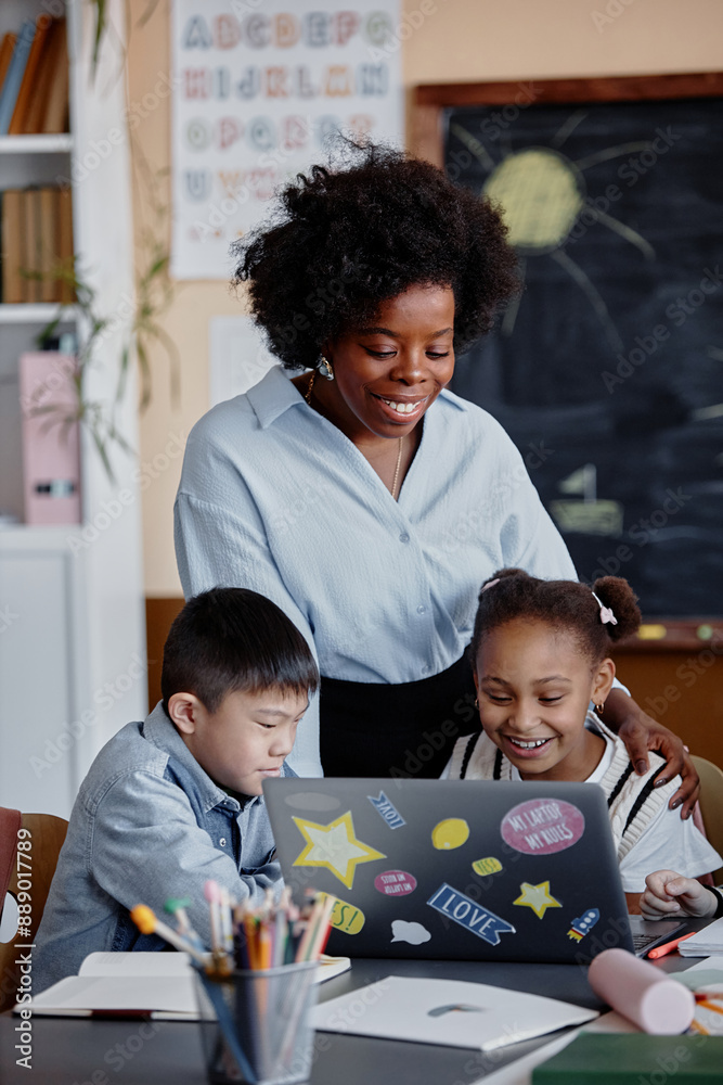 © Seventyfour - Vertical shot of smiling female teacher of Black ethnicity talking to primary students looking at laptop screen