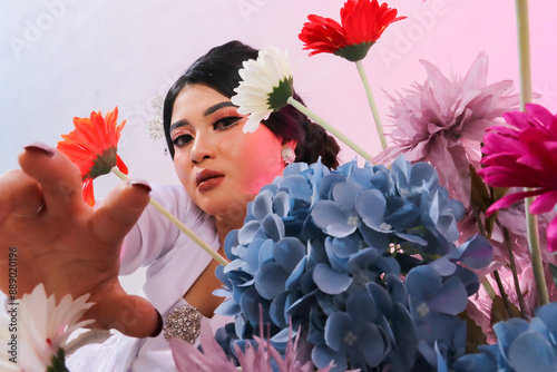 closeup of beautiful young elegant Asian woman standing next to flower decorations wearing modern Javanese traditional clothing, white velvet bridal kebaya on a white studio background photo