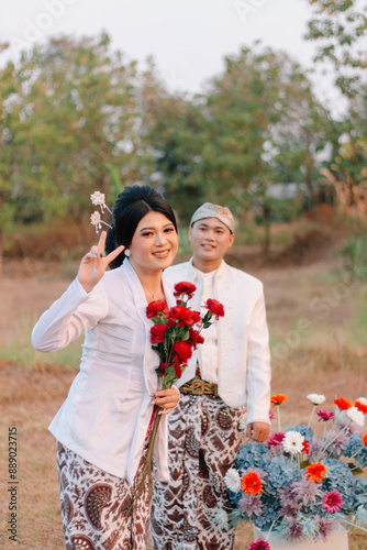 A pair of adult Asian men and woman lovers standing, the woman smiling peacefully in front of the man wearing modern Javanese traditional clothing, the bride's white velvet in an outdoor barren photo