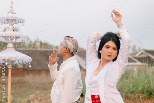 couple young Asian man and woman lovers standing, elegant woman behind the man smoking wearing modern Javanese traditional clothing, white velvet for the bride and groom in an outdoor barren photo